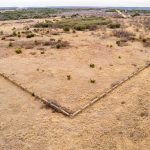 Aerial view of the Historic S&S Ranch shows a dry, grassy field with a V-shaped fence enclosing land. Sparse shrubs and bushes dot the landscape, set against distant rolling hills and cloudy skies.