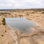 A small pond at the historic S&S Ranch is surrounded by dry, brown grass and sparse bushes in a wide, arid landscape with distant hills under a cloudy sky. Dirt roads run alongside the pond and through the scene.