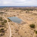 Aerial view of the dry, sparsely vegetated landscape of Historic S&S Ranch, featuring a small, irregularly shaped pond near dirt roads and patches of shrubs and trees under a cloudy sky.