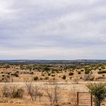 A wide, dry landscape with sparse bushes and small trees under a cloudy sky. The ground is mostly yellowish-brown, and a rusty gate marks the entrance to the historic S&S Ranch, with distant hills lining the horizon.