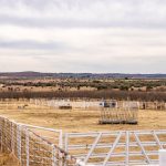 A wide, open landscape with dry grass, scattered bushes, and distant hills surrounds the Historic S&S Ranch. White metal fencing encloses a large, empty corral area under a mostly cloudy sky.