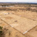 Aerial view of the Historic S&S Ranch shows a large, rectangular, fenced enclosure on dry, grassy land with scattered shrubs, dirt paths, and a small distant pond in a rural landscape.