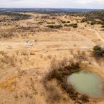Aerial view of S&S Ranch 1, a dry, grassy landscape with sparse shrubs and trees, a small green pond in the foreground, dirt roads, and fenced areas. Distant rolling hills are visible under a cloudy sky on this historic ranch.