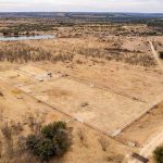 An aerial view of the Historic S&S Ranch reveals a dry, rural landscape with a fenced rectangular area, dirt roads, sparse trees, and a small pond in the background under an overcast sky.