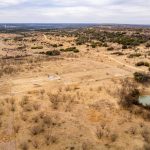 Aerial view of the Historic S&S Ranch, showcasing a dry, grassy landscape with scattered bushes, a small pond, dirt roads, and fencing. Rolling hills and cloudy skies form a scenic backdrop.