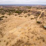Aerial view of the Historic S&S Ranch reveals a vast, dry landscape with sparse vegetation, dirt paths, a small pond, and rectangular foundations near the center. Rolling hills extend into the distance.