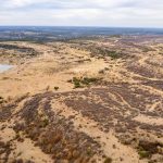 A wide aerial view of S&S Ranch shows a dry, rolling landscape with sparse vegetation, patches of brush, a small pond on the left, and distant hills under a partly cloudy sky.