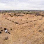 Aerial view of the Historic S&S Ranch shows a dry, open field with cattle gathered near a white car and a person standing nearby. An enclosed rectangular area with a fence sits in the center of this expansive landscape.