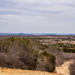 A wide view of the dry, open landscape at Historic S&S Ranch, with sparse trees and bushes under a cloudy sky. Rolling hills and distant mountains stretch across the horizon, while a dirt path and utility pole sit on the right side.