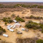 Aerial view of the Historic S&S Ranch campsite, with several RVs and trailers scattered around a central building, surrounded by dry grass, bushes, a small pond, and dirt roads winding through scenic hills in the background.
