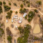 Aerial view of the Historic S&S Ranch campsite with several RVs, buildings, clustered trees, dirt roads, and a small muddy pond on dry, brown grassland. Vehicles are parked throughout the site.