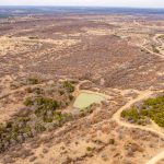 Aerial view of the semi-arid landscape at Historic S&S Ranch, with patches of trees, winding dirt roads, and a small pond near the center. The dry, sparsely vegetated land stretches toward the horizon.