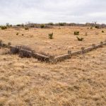A V-shaped stone wall at the Historic S&S Ranch stands in a dry, grassy field under a cloudy sky, with scattered bushes and sparse vegetation in the background.