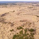Aerial view of the Historic S&S Ranch reveals a dry, open landscape with patches of shrubs and grass, dirt roads, and scattered small structures. The terrain is mostly flat with gentle hills in the distance under a clear sky.