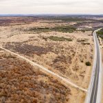 Aerial view of a rural landscape with a winding paved road cutting through dry, sparsely vegetated land and patches of shrubs beneath a cloudy sky, near the Historic S&S Ranch.