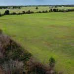 Aerial view of a large, open grassy field bordered by scattered trees and shrubs, with a road and distant buildings visible in the background under a partly cloudy sky.