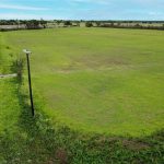A wide, open grassy field sits beside a two-lane rural road. A white truck drives along the road, and a utility pole stands near the edge of the field. The sky is clear with a few clouds.