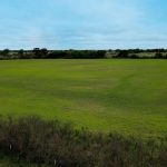 A wide, open grassy field with patches of green and brown, bordered by trees and bushes under a cloudy blue sky. A line of fence posts runs along the edge of the field in the foreground.