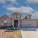 A single-story brick house with a two-car garage, front porch, and neatly landscaped yard under a partly cloudy blue sky.