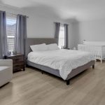 A modern, minimal bedroom with a gray upholstered bed, white bedding, a white crib, a white armchair, two windows with gray curtains, and light wood flooring. A small shelf holds baby items near the crib.