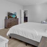 A modern bedroom with a white crib, gray bed with white bedding, wooden dresser with a TV, armchair near a window with gray curtains, and light wood flooring. Multiple open doors lead to adjoining rooms.