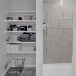 Open shelving in a bathroom with towels, toilet paper rolls, toiletries, and baskets neatly arranged; a glass scale and wire laundry basket on the floor, next to a shower with light beige tiles and a white curtain.