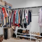 A walk-in closet with neatly hung shirts, jackets, and pants. Shelves below hold shoes and boots. Storage bins and folded blankets are on the upper shelf, and the closet has light-colored walls and flooring.