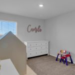 A small, bright children's room with a white dresser, a toy house, stuffed animals, a play table with chairs, and “Camila” in pink letters on the wall above the dresser. A window lets in natural light.