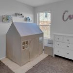 A children's bedroom with a play tent shaped like a house, a white dresser, pink storage bins, plush toys, wall shelves with books, and the name "Camila" in cursive letters above the dresser.