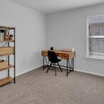 Minimalist home office with a wooden desk, black chair, pink typewriter, and bookshelf with baskets and books on beige carpet, next to a window with white blinds and gray walls.