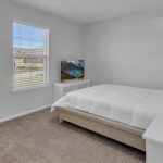 A simple bedroom with a bed covered in white bedding, a small TV on a stand, a window with blinds letting in natural light, a white nightstand, and a laundry basket in the corner on carpeted flooring.