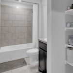 A modern bathroom with a tiled bathtub and shower, white shower curtain, toilet, dark vanity with a sink, and open shelves stocked with towels, toilet paper, and toiletries.