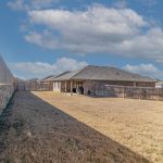 A large backyard with dry grass, bordered by a tall wooden fence on the right and a stone wall on the left, leads to a single-story brick house under a partly cloudy sky.