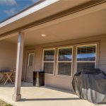 A covered backyard patio with a small table and two chairs, a grill with a cover, and a mini fridge against a beige house with multiple windows and a wooden fence in the background.