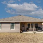 A single-story house with taupe siding, three windows, and a covered patio. The backyard has dry grass, a wooden fence, a grill, and a small table with a chair. The sky is mostly clear with a few clouds.