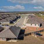 Aerial view of a suburban neighborhood with rows of houses, fenced backyards, a trampoline, a patio, and several parked cars along the streets under a partly cloudy sky.