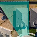 Aerial view of a suburban home with a triangular shaded area covering the backyard and part of the roof, surrounded by neighboring houses, fences, and a circular trampoline.