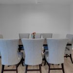 A minimalist dining room with a dark rectangular table, eight light gray upholstered chairs, and a small vase with red flowers at the center. The room has light wood flooring and plain gray walls.