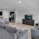 A modern open-concept living room and kitchen with gray sofas, a TV on a dark cabinet, a breakfast bar with two chairs, stainless steel appliances, and light wooden floors. Large windows let in natural light.