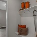 A small entryway with a white bench, orange and brown pillows, a clear umbrella, and a shelf holding baskets and vases. The space leads to a bright room with a large window and wooden floor.