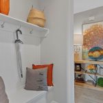 A cozy entryway with a white bench, orange and gray pillows, a hanging umbrella, and wall hooks. In the background, a console table with lamps, decorative items, and a colorful painting is visible.