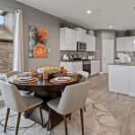 A modern kitchen and dining area with light gray walls, wood flooring, a round dining table set for four, white cabinets, stainless steel appliances, and a large window letting in natural light.