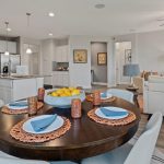 Open-concept kitchen, dining, and living area with a round dining table set for four, modern white kitchen cabinets, light gray walls, and a beige sofa facing a TV in the living room.