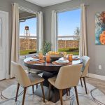 A modern dining area with a round table set for four, beige chairs, orange décor accents, a fruit bowl centerpiece, large windows with beige curtains, wall art, and a view of a brick fence and water tower outside.