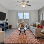 A modern living room with beige sofas, colorful accent pillows, a glass coffee table on an orange geometric rug, a wall-mounted TV, and large windows letting in natural light.