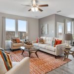 Bright living room with light-colored sofas, orange and striped accent pillows, an orange geometric rug, a glass coffee table, and large windows letting in natural light. Dining area is visible in the background.