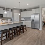 Modern kitchen with white cabinets, stainless steel appliances, gray countertops, and a large island with three dark wooden stools. Pendant lights hang above the island, and the kitchen opens to a hallway with wood-look flooring.