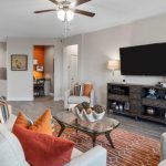 A modern living room with beige sofas, orange and patterned cushions, a glass coffee table, and a TV on a dark console. An orange rug and decor accent the space. A kitchen and small office area are visible in the background.