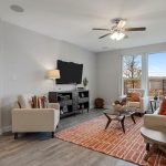 A modern living room with light gray walls, wood flooring, a beige sofa, two armchairs, a wall-mounted TV, a ceiling fan, large windows, and an orange patterned rug with matching pillows.