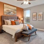 A modern bedroom with a wooden bed, white bedding, orange and gray pillows, two bedside tables with lamps, a bench at the foot of the bed, large windows with sheer curtains, and wall art above the bed.